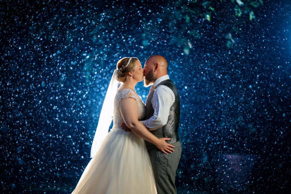 Ein Brautpaar steht bei einer Hochzeit mit schlechtem Wetter verliebt im Regen und küsst sich in einem Foto von einem professionellen Hochzeitsfotografen 